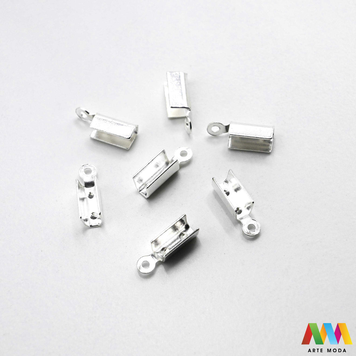 Metalni stiskači i završeci sa zubima za izradu nakita u srebrnoj, zlatnoj i brončanoj boji složeni na radnoj površini. Prikazani su ravni stiskači za tekstilne trake, baršun, antilop i plosnate kožice s integriranom omčicom za spajanje s kopčama i produžnim lančićima. Kvalitetan repromaterijal za uredno fiksiranje krajeva pri izradi choker ogrlica, narukvica i modnih dodataka u DIY radionicama za ručnu izradu unikatnog nakita s nazubljenim rubovima koji jamče čvrst prihvat bez ispadanja materijala.