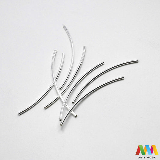 Sjajne metalne zakrivljene cijevčice i noodle distanceri u srebrnoj i zlatnoj boji složeni na radnoj površini za izradu nakita. Prikazane su glatke i gravirane savijene metalne perle u obliku cijevi, namijenjene za provlačenje konca, gume, flaksa ili kožice pri izradi unikatnih narukvica i ogrlica. Vrhunski metalni repromaterijal i razdjelnici za DIY izradu modernog geometrijskog i elegantnog ručno rađenog nakita s precizno izbušenim otvorima za lako nizanje perli i kristala u kreativnim radionicama.